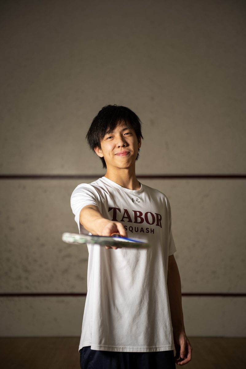 Jimmy on a squash court in a Tabor Squash shirt, holding a racket toward the camera